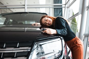 White-appearing woman with red hair wearing black shirt and orange pants smiling and hugging her new black SUV.